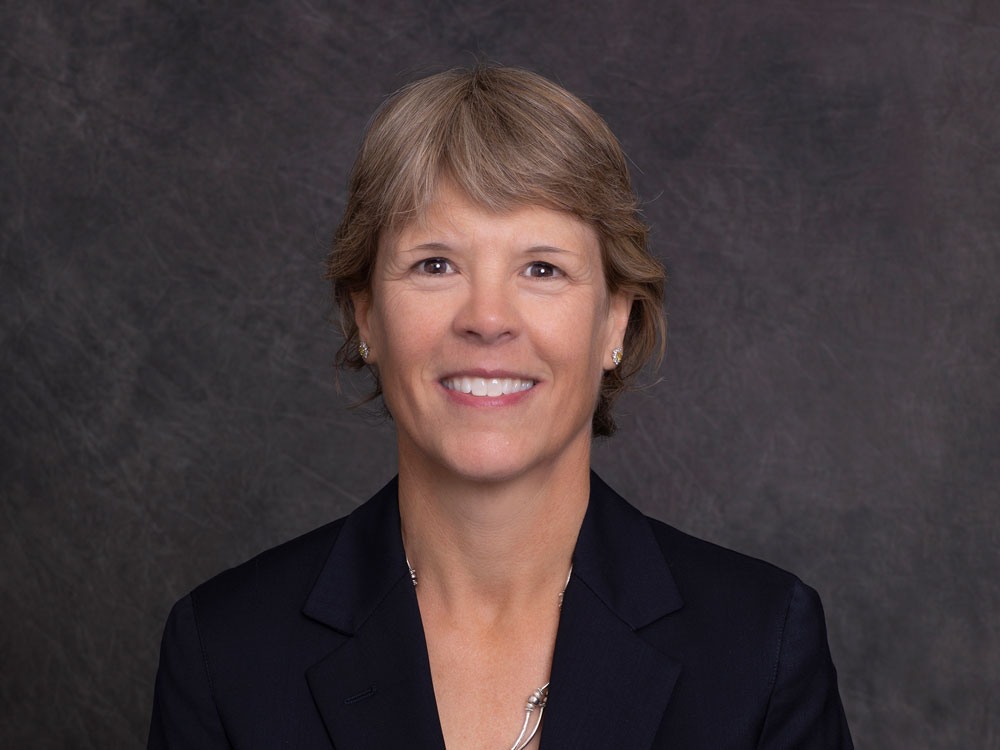 Head and shoulders portrait of Susan Gellatly, a white woman with a toothy smile and dark blond hair cropped short, dressed in professional attire